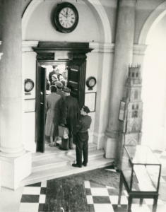 black and white photo. people queueing to enter tall doors in an arched doorway. Floor is black and white tiles and there is a glass display case.