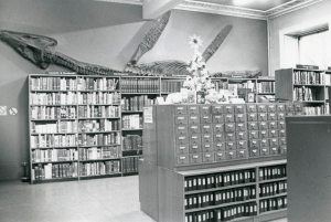 black and white photo, card catalogue in foreground, bookshelves against wall with large dinosaur fossil-type mural on wall
