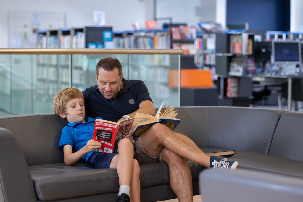 man and boy sat on sofa reading in front of glass balustrade. Full bookshelves in background.