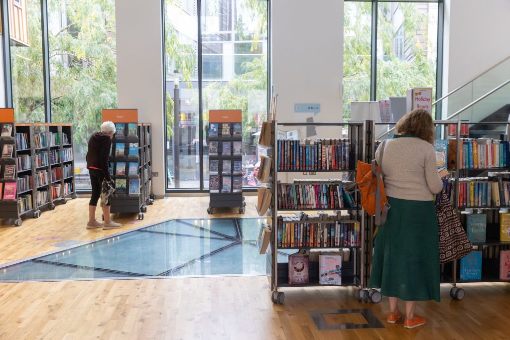 open space with people browsing full bookshelves. double-height full length windows in background.