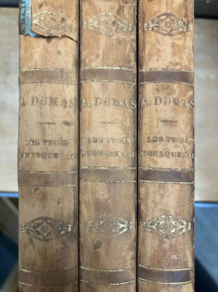 Spines of 3 copies of Les Trois Mousquetaires. faded and slightly torn beige, with embossed line decorations.