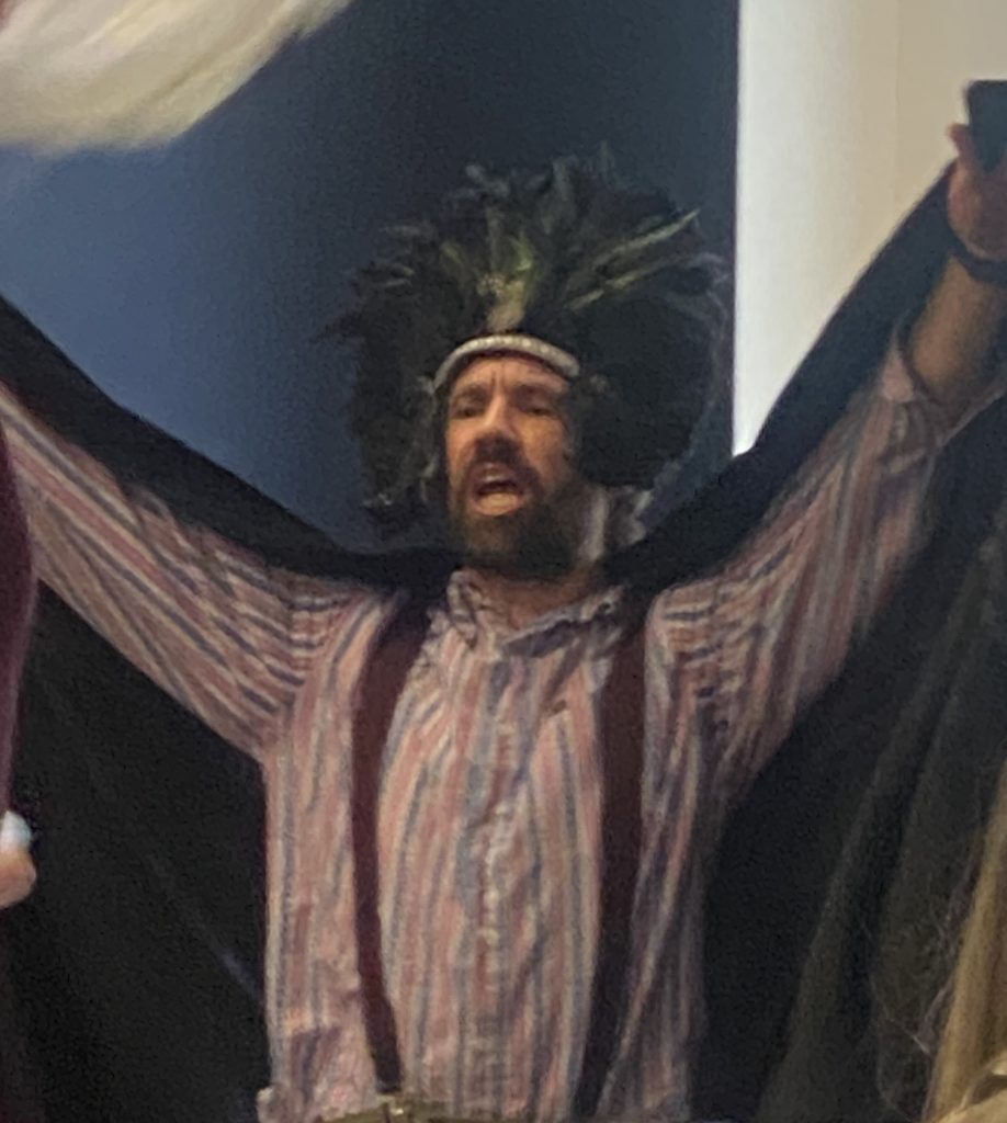 Man in feathered headdress and cape with arms outstretched