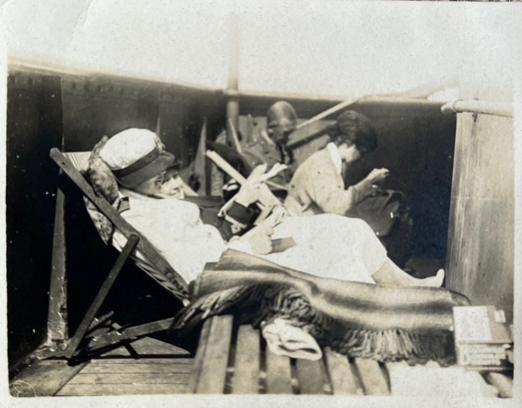 black and white photograph of woman in hat and long white dress on a deck chair reading. a woman beside her smiles at the camera.