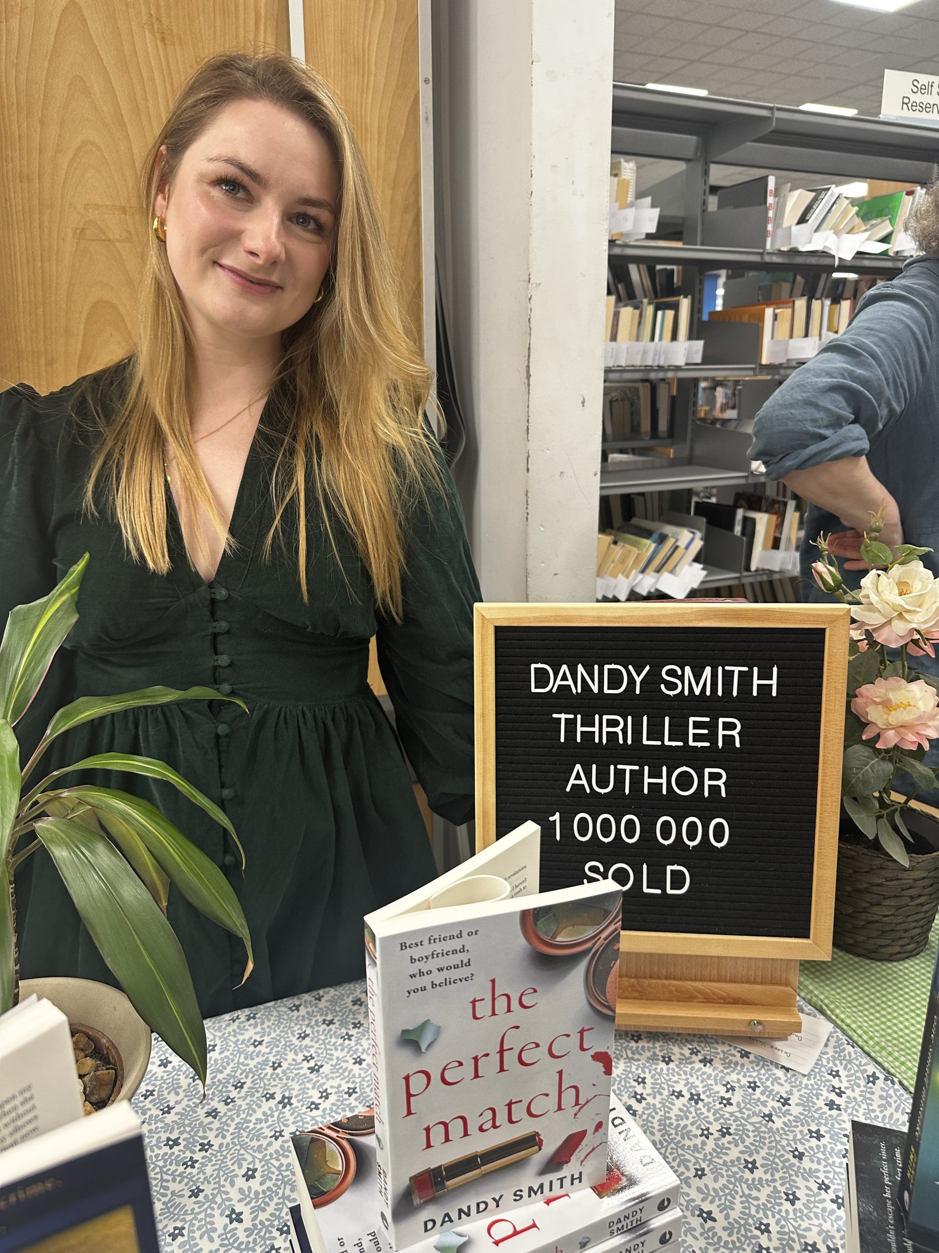 woman smiling next to pegboard sign saying 'Dandy Smith Thriller Author 1 000 000 Sold'