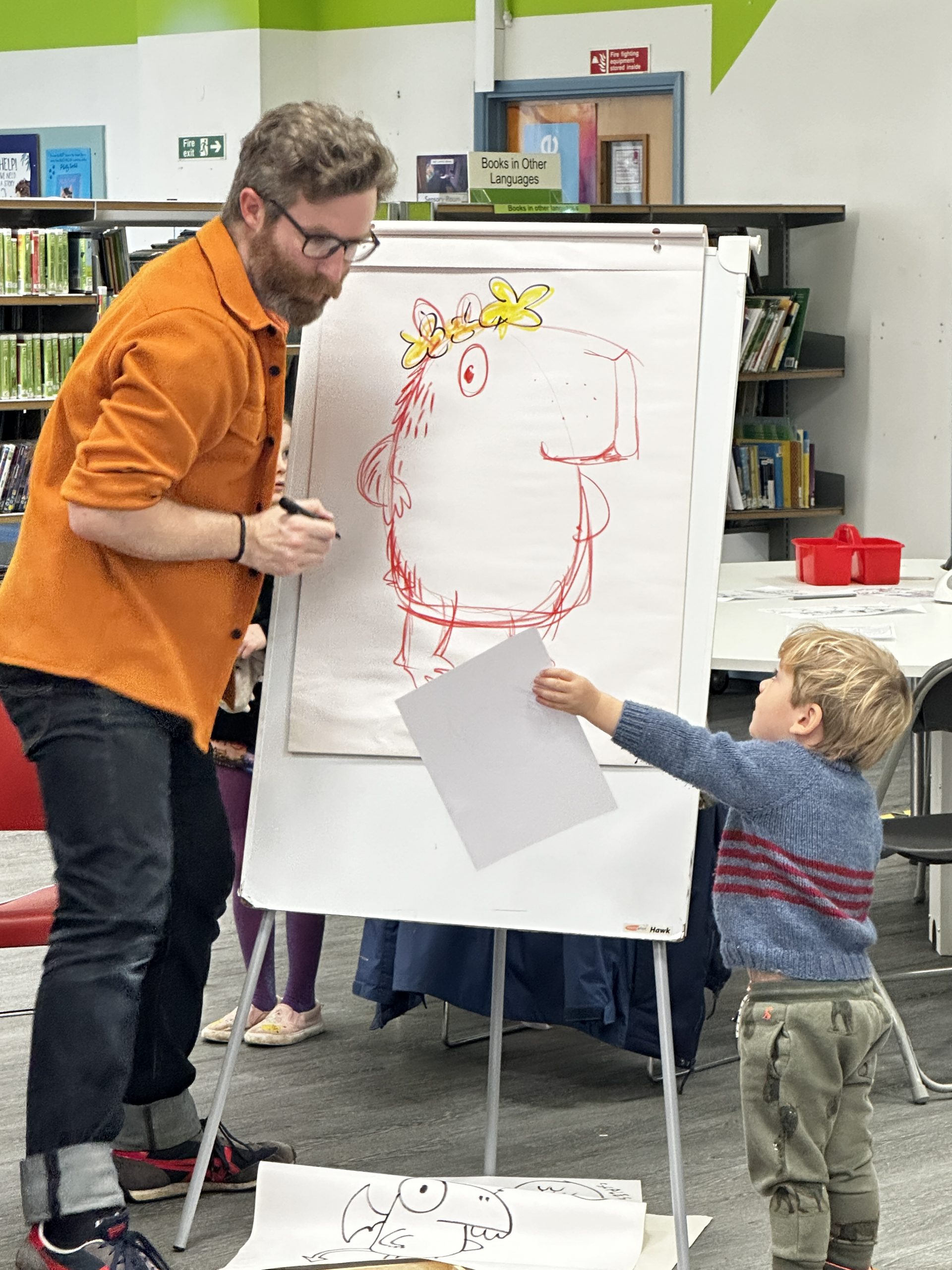 man stood at flipchart with drawing of creature looking at a piece of paper held up by a little boy
