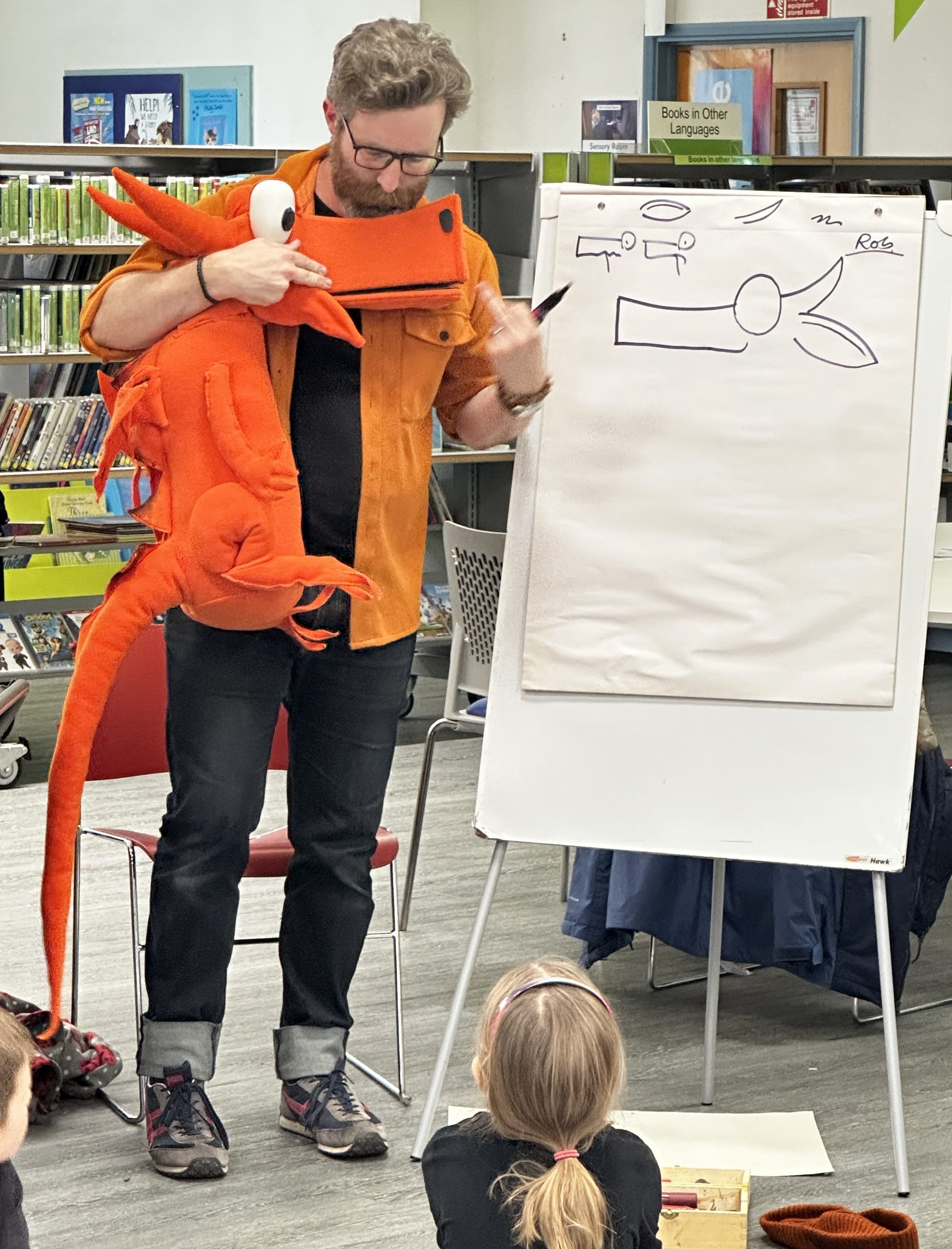 Man holding large orange toy dragon and pen next to flip chart