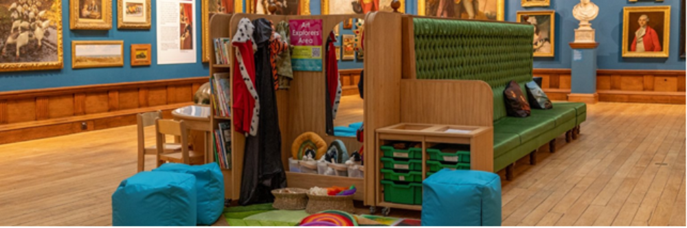 children's area, with bean bags, dress up clothes, small table with chairs and books in the middle of a wood panelled art gallery